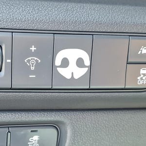 May include: A car dashboard with various buttons. The buttons are dark grey with white symbols. One button displays a white image of a dog's nose. Other buttons show symbols for lights and windscreen wipers.