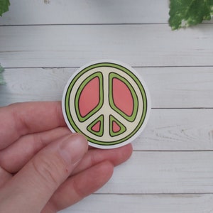 Pu&ograve; includere: Un adesivo rotondo con un design a forma di segno della pace. Il segno della pace &egrave; contornato di verde e riempito con un colore rosa chiaro. L'adesivo &egrave; su uno sfondo di legno bianco.