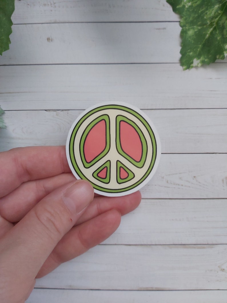 Pu&ograve; includere: Un adesivo rotondo con un design a forma di segno della pace. Il segno della pace &egrave; contornato di verde e riempito con un colore rosa chiaro. L'adesivo &egrave; su uno sfondo di legno bianco.