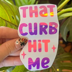 Può includere: Un adesivo bianco con uno sfondo di tramonto rosa, viola e arancione. L'adesivo dice "That Curb Hit Me" in un font a bolle.
