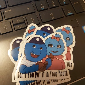 May include: Three blue fuzzy monster stickers with a white background. The stickers have a blue and white striped shirt, a pink dress, and a blue hat. The text on the stickers reads "I Don't You Put it in Your Mouth Don't You Put it in Your Mouth".
