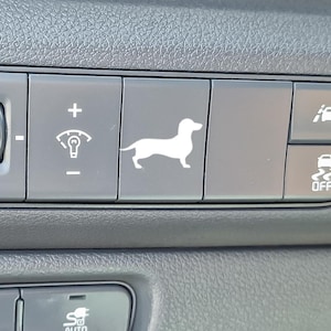 May include: Close-up of a car dashboard with several buttons. One button features a white silhouette of a dachshund dog. Other buttons include symbols for lights and other car functions. The dashboard is dark gray with a textured surface.