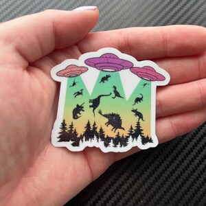 May include: A sticker with a rainbow gradient background featuring a group of dinosaurs being abducted by three flying saucers. The sticker is on a black and white carbon fiber background.