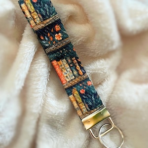 May include: A dark green and orange key chain with a bookshelf design and a silver key ring. The key chain features a pattern of books, flowers, and leaves.