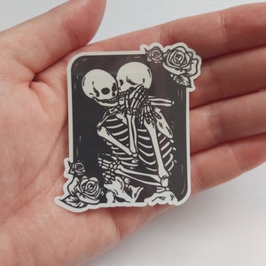 May include: A black and white sticker featuring a couple of skeletons embracing and kissing. The skeletons are surrounded by a black frame with white roses at the top and bottom.