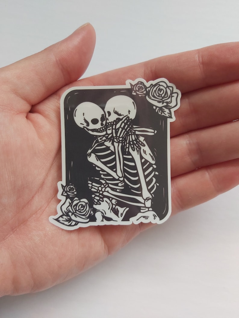 May include: A black and white sticker featuring a couple of skeletons embracing and kissing. The skeletons are surrounded by a black frame with white roses at the top and bottom.