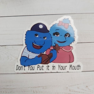 May include: A sticker featuring two blue fuzzy characters, one wearing a blue and white striped shirt and a blue hat, the other wearing a pink shirt and a pink bow. The characters are holding a brown chocolate bar and the text "Don't You Put it in Your Mouth" is below them.