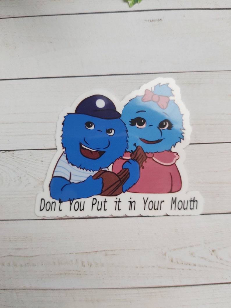 May include: A sticker featuring two blue fuzzy characters, one wearing a blue and white striped shirt and a blue hat, the other wearing a pink shirt and a pink bow. The characters are holding a brown chocolate bar and the text "Don't You Put it in Your Mouth" is below them.