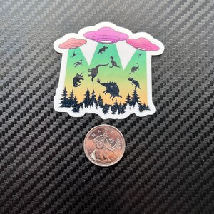 May include: A sticker featuring a group of dinosaurs being abducted by three flying saucers. The dinosaurs are silhouetted against a forest background with a gradient of green, yellow, orange, and red.