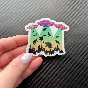 May include: A sticker with a rainbow gradient background featuring a silhouette of a forest with dinosaurs being abducted by three flying saucers. The saucers are pink, purple, and blue.