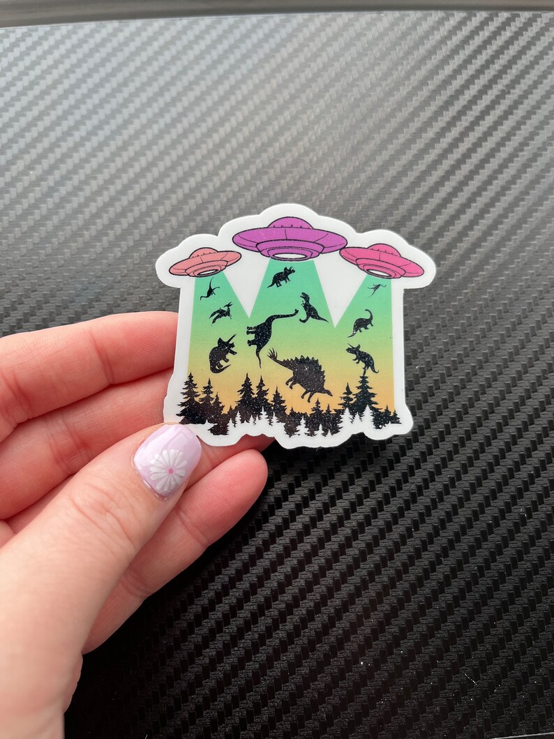 May include: A sticker with a rainbow gradient background featuring a silhouette of a forest with dinosaurs being abducted by three flying saucers. The saucers are pink, purple, and blue.