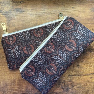 Peut inclure: Deux trousses rectangulaires à fermeture éclair avec un motif en tissu sombre représentant des petits dragons, des vignes entrelacées et des feuilles claires. Chaque trousse possède une fermeture éclair blanche et une tirette dorée.