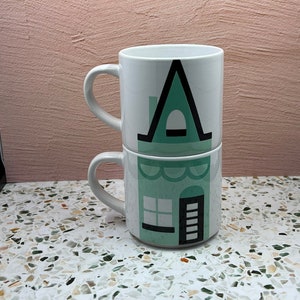 May include: Two white ceramic mugs stacked on top of each other. The mugs feature a green and black illustration of a house with a window and door.