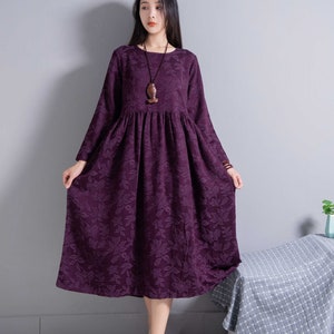 May include: A long-sleeved, purple dress with a floral pattern. The dress has a round neckline and a gathered waist. The model is wearing a wooden pendant necklace.