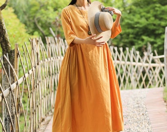 May include: A woman wearing a yellow linen dress and a straw hat with a black and white checkered ribbon. She is standing on a path in a garden.