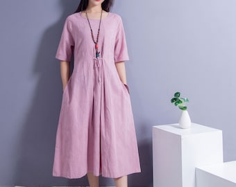 May include: A woman wearing a pink linen dress with a beaded necklace. She is standing in front of a light gray wall with white platforms behind her.