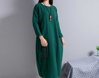 May include: A woman wearing a long, green, textured dress with a wooden fish pendant necklace. She is standing in front of a gray wall and a checkered tablecloth.