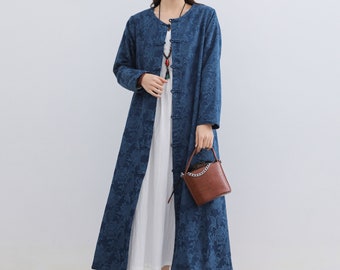 May include: A woman wearing a long, dark blue floral patterned coat over a white dress. The coat has a mandarin collar and frog closures. She is holding a brown handbag and wearing white shoes. A woven wall decoration is in the background.