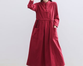 May include: A long-sleeved, red, button-up dress with a mandarin collar and two pockets. The dress has a cinched waist and a flowing skirt. The model is wearing a straw hat.