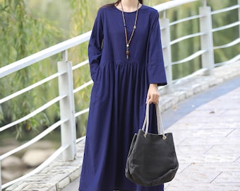 May include: A woman wearing a navy blue long-sleeved dress with a round neckline and a gathered skirt. She is carrying a dark gray tote bag with white handles and wearing a light gray hat.