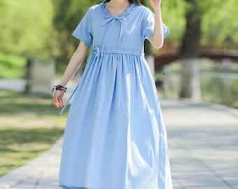 May include: A light blue midi dress with a ruffled neckline and a bow tie. The dress has short sleeves and a gathered skirt. The model is wearing a straw hat and white sandals.