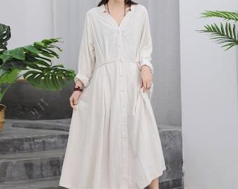 May include: A long-sleeved, button-down dress in a light beige color. The dress features a V-neckline, a cinched waist, and a flowing skirt. A black bucket hat and white slip-on shoes complete the outfit. The dress is made of a lightweight fabric.