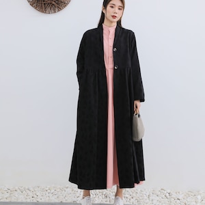 May include: A black long-sleeved coat with a button closure and a slightly flared hem. The coat is worn over a pink dress. The model is wearing white shoes.