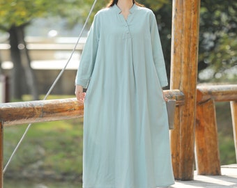 May include: A light blue long-sleeved maxi dress with a V-neckline and a single button closure. The dress is made of a lightweight fabric and has a relaxed fit. The model is wearing a straw hat and standing on a wooden bridge.