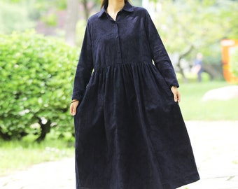 May include: A woman wearing a long, dark blue linen dress with a button-up front and long sleeves. She is wearing a straw hat and standing on a stone path.