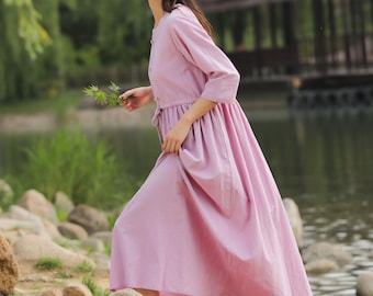 May include: A woman in a lavender-colored dress stands by a pond. She is holding a small branch with green leaves. The dress has a cinched waist and a flowing skirt.