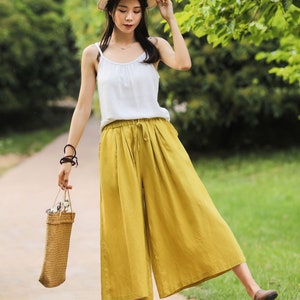 May include: A woman wearing a white tank top and yellow wide-leg pants with a straw hat and a woven bag.