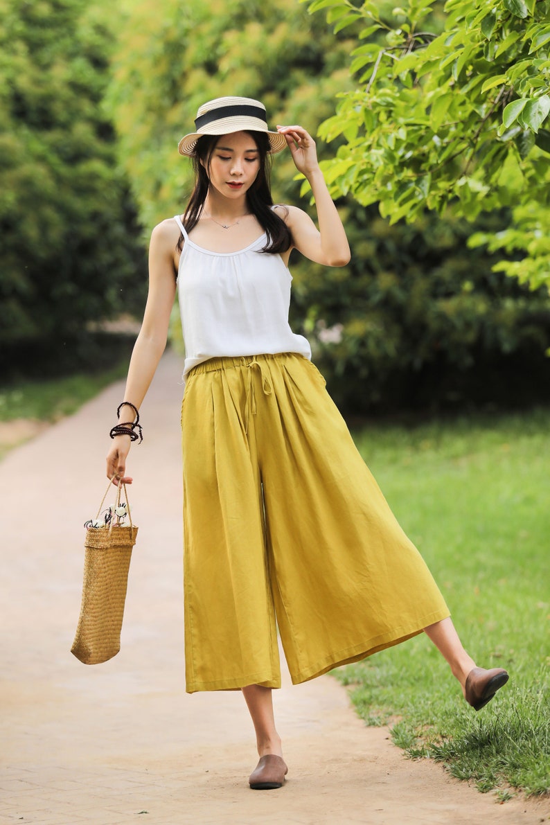 May include: A woman wearing a white tank top and yellow wide-leg pants with a straw hat and a woven bag.
