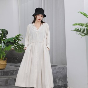 Puede incluir: Un vestido largo color crema con mangas largas y botones en la parte delantera. El vestido presenta un escote en V, una cintura ce&ntilde;ida y una falda fluida. Un sombrero de pescador negro se usa con el vestido. El vestido se combina con zapatos blancos.