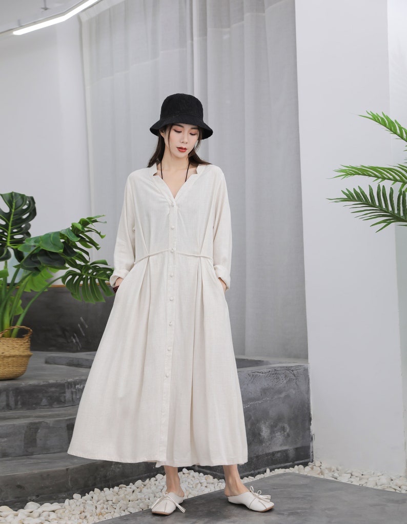 Puede incluir: Un vestido largo color crema con mangas largas y botones en la parte delantera. El vestido presenta un escote en V, una cintura ce&ntilde;ida y una falda fluida. Un sombrero de pescador negro se usa con el vestido. El vestido se combina con zapatos blancos.