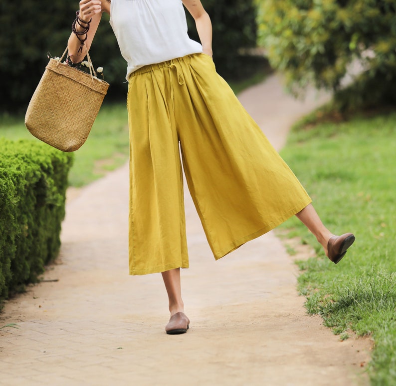 May include: A woman wearing a white tank top and mustard yellow wide-leg culottes. She is carrying a woven straw bag and is walking on a paved path.