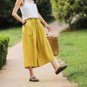 May include: A woman wearing a white tank top and mustard yellow wide-leg linen pants. She is carrying a woven straw bag with white flowers.
