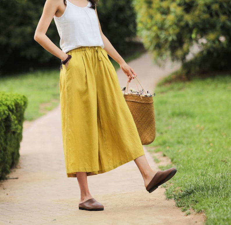 May include: A woman wearing a white tank top and mustard yellow wide-leg linen pants. She is carrying a woven straw bag with white flowers.