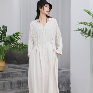 Puede incluir: Una mujer con un vestido largo de lino color crema con cuello en V y botones. El vestido tiene mangas largas y cintura ce&ntilde;ida. Tambi&eacute;n lleva un sombrero de pescador negro y zapatos blancos.