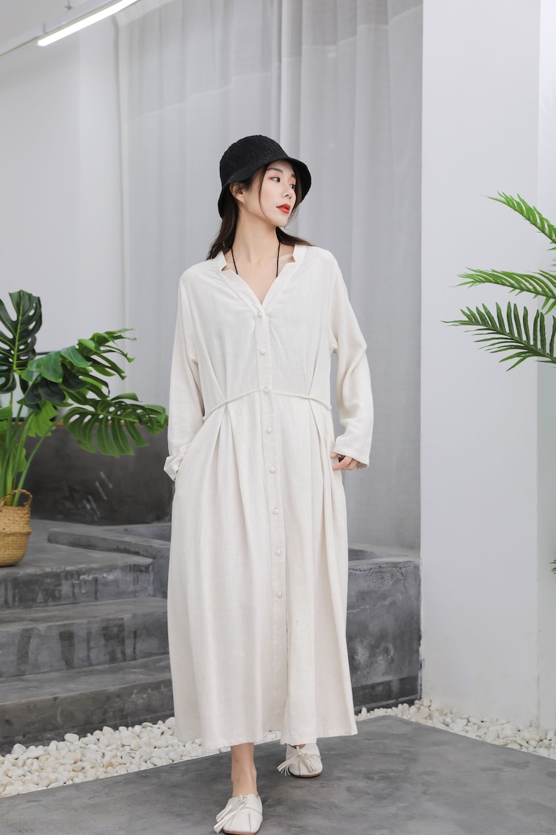 Puede incluir: Una mujer con un vestido largo de lino color crema con cuello en V y botones. El vestido tiene mangas largas y cintura ce&ntilde;ida. Tambi&eacute;n lleva un sombrero de pescador negro y zapatos blancos.