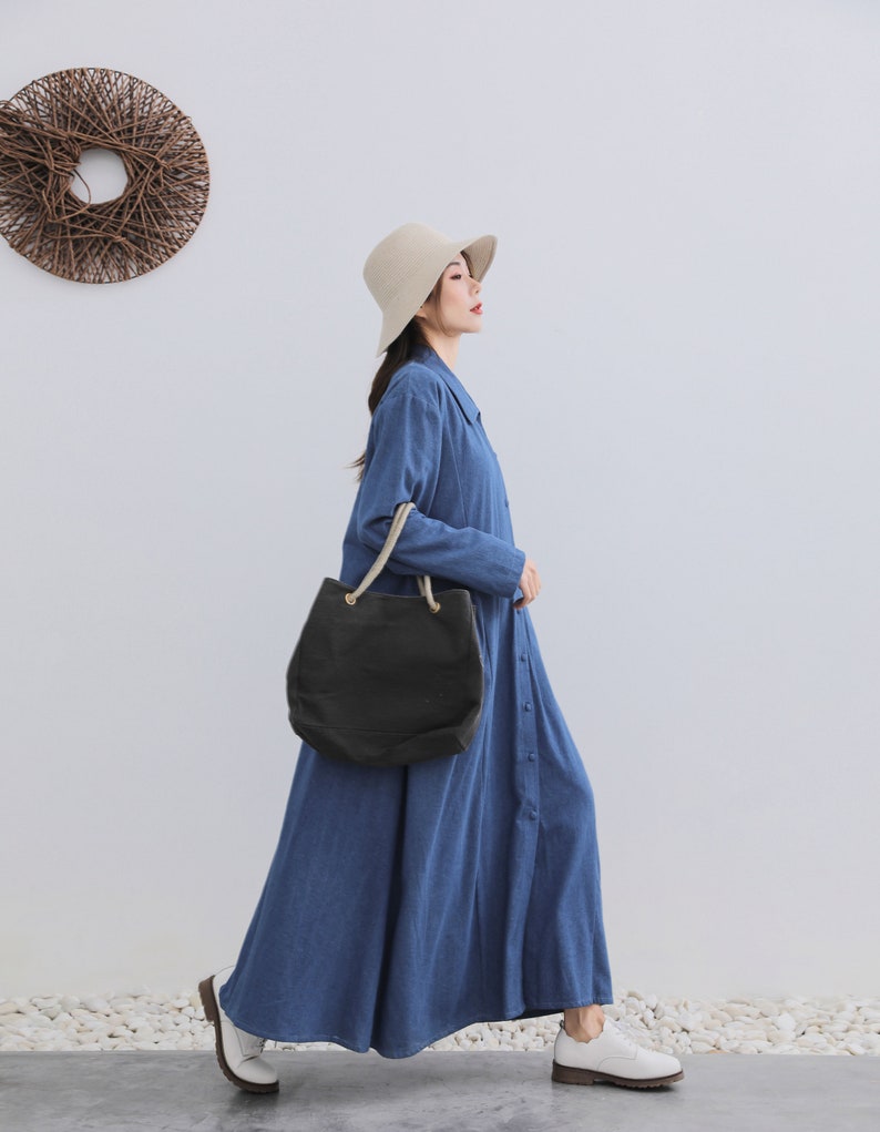 May include: A woman wearing a long, blue, button-down dress with a black tote bag and a white hat. She is walking on a white surface.
