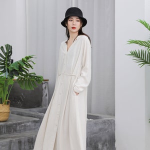 Puede incluir: Vestido largo color crema con botones, mangas largas y cintur&oacute;n en la cintura. El vestido se combina con un sombrero de pescador negro y zapatos blancos. El vestido tiene bolsillos y una falda fluida.