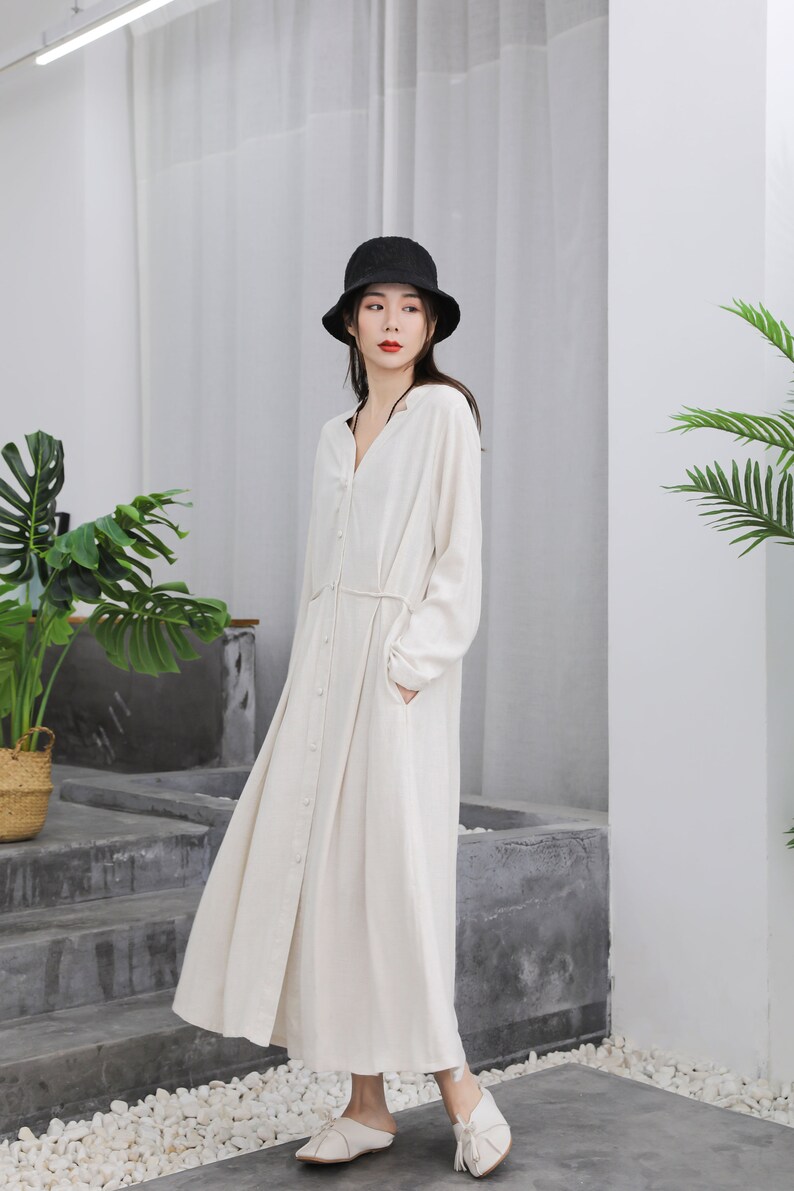 Puede incluir: Vestido largo color crema con botones, mangas largas y cintur&oacute;n en la cintura. El vestido se combina con un sombrero de pescador negro y zapatos blancos. El vestido tiene bolsillos y una falda fluida.