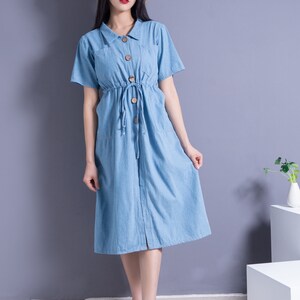 May include: A woman wearing a light blue denim dress with a button-up front and a drawstring waist. She is wearing a straw hat and white shoes.