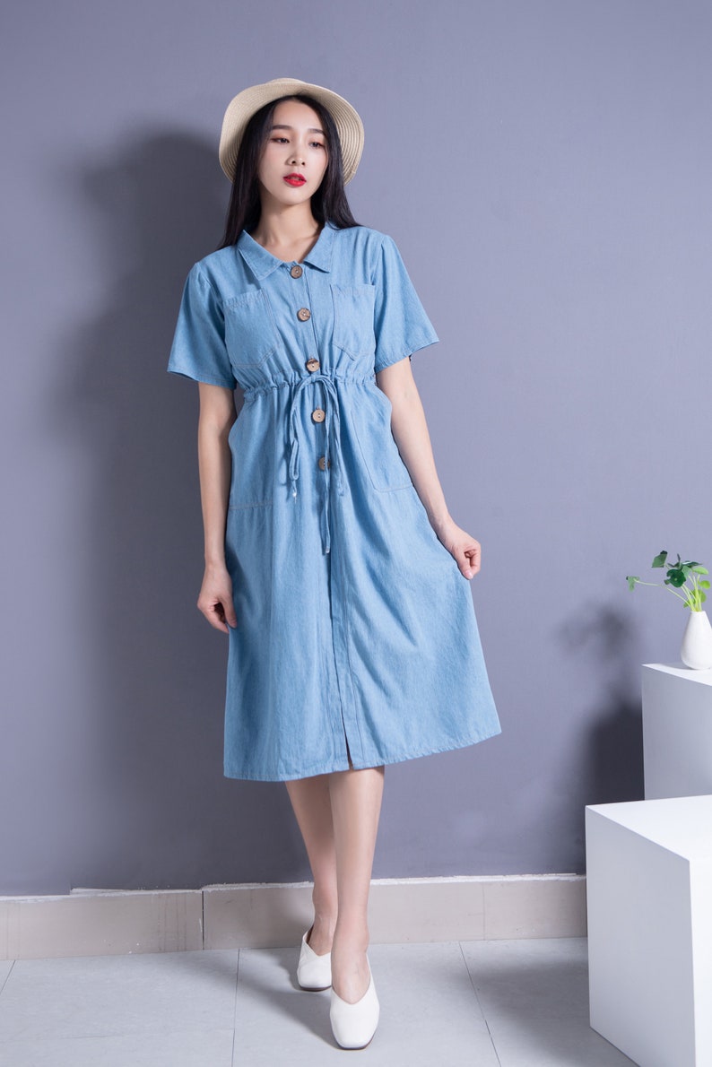 May include: A woman wearing a light blue denim dress with a button-up front and a drawstring waist. She is wearing a straw hat and white shoes.