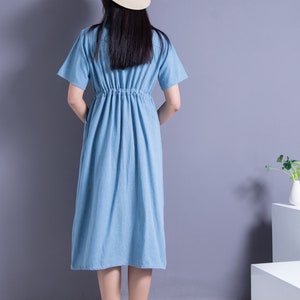 May include: A light blue denim dress with a cinched waist and short sleeves. The dress is worn by a person wearing a straw hat with a black ribbon.