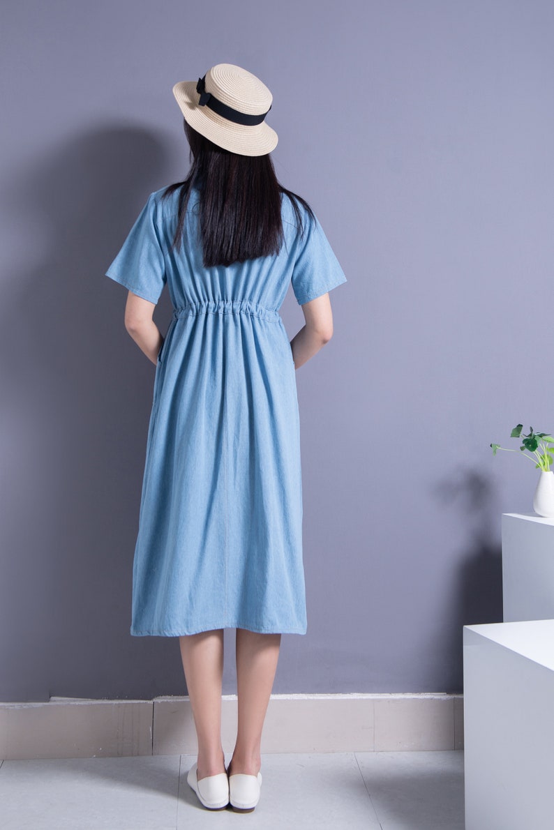 May include: A light blue denim dress with a cinched waist and short sleeves. The dress is worn by a person wearing a straw hat with a black ribbon.