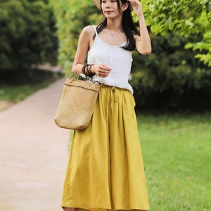 May include: A woman wearing a white tank top, a straw hat, and yellow wide-leg pants. She is carrying a woven tote bag and is walking on a path.