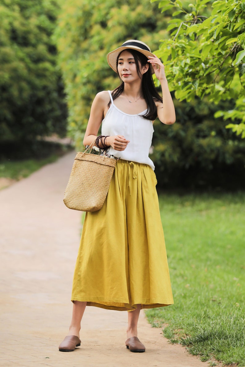May include: A woman wearing a white tank top, a straw hat, and yellow wide-leg pants. She is carrying a woven tote bag and is walking on a path.
