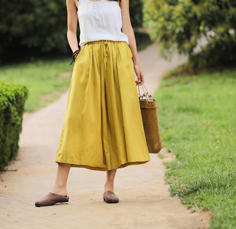 May include: A woman wearing a white tank top and mustard yellow wide-leg culottes. She is carrying a woven straw bag.