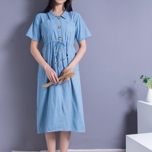 May include: A woman wearing a light blue denim dress with a straw hat and white shoes. She is holding a bunch of dried wheat stalks.